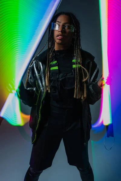 Elegante mujer afroamericana en gafas inteligentes posando con palos de neón sobre fondo gris - foto de stock