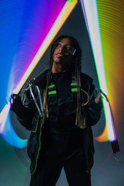 Fashionable african american woman in smart glasses holding abstracted neon sticks on grey background — Stock Photo