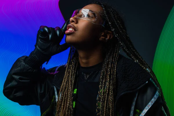 Mujer afroamericana en guante y gafas inteligentes mirando hacia otro lado cerca de la abstracción colorida sobre fondo gris - foto de stock