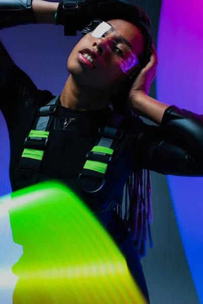 Elegante donna afro-americana in occhiali intelligenti in posa vicino a luci al neon su sfondo colorato — Foto stock