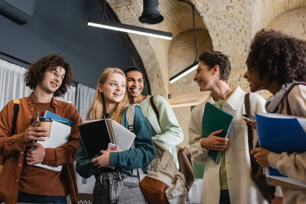 Gruppe junger interrassischer Studenten mit Notizbüchern und Gadgets, die sich in der Universität anschauen — Stockfoto