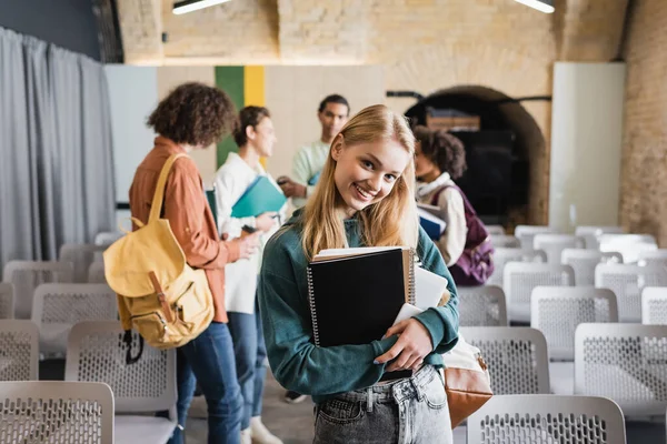 Pretty student with copybooks smiling at camera near interracial classmates on blurred background — Stock Photo