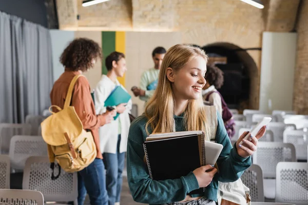 Femme souriante avec des cahiers en utilisant smartphone proches amis multiethniques dans l'auditorium flou — Photo de stock