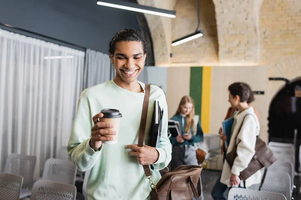 Sonriente hombre afroamericano con bolsa de cuero, copybooks y taza de papel cerca de compañeros de clase borrosos - foto de stock