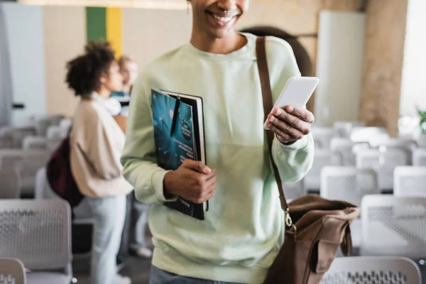 Vista recortada del hombre afroamericano con revista y cuaderno usando teléfono inteligente sobre fondo borroso - foto de stock