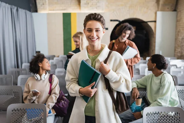 Mujer alegre con copybooks mirando la cámara cerca de los estudiantes multiétnicos en el auditorio borroso - foto de stock