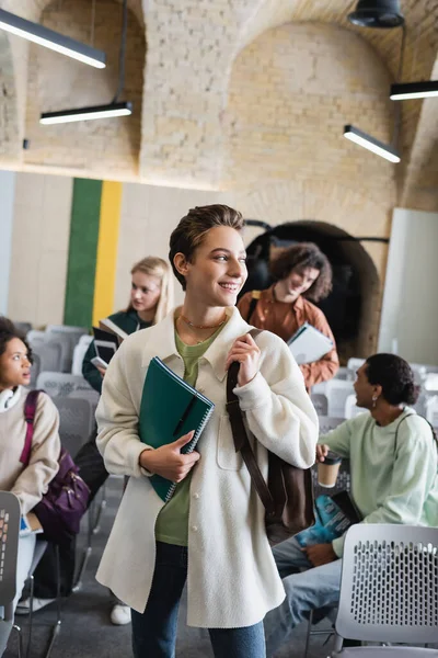 Femme heureuse avec des copybooks regardant loin près flous amis interracial dans la salle de classe — Photo de stock