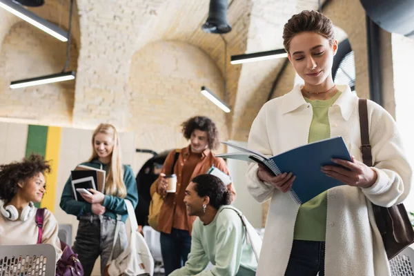 Junge Frau schaut in Werbebuch in der Nähe multiethnischer Studenten mit Gadgets auf verschwommenem Hintergrund — Stockfoto