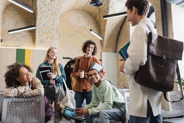 Camarades de classe interracial joyeux avec des gadgets et des cahiers regardant étudiant avec sac à dos en cuir sur le premier plan flou — Photo de stock