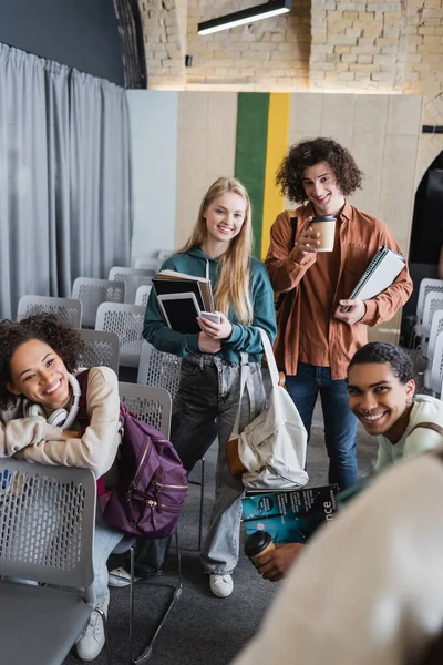 Jóvenes estudiantes interracial con papeles tazas, gadgets y cuadernos sonriendo a la cámara — Stock Photo