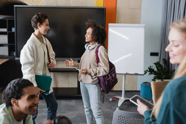 Heureux étudiant afro-américain avec sac à dos parler à un ami près de camarades de classe multiethniques — Photo de stock