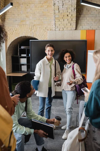 Heureux interracial femmes souriant près des étudiants et tableau à l'université — Photo de stock