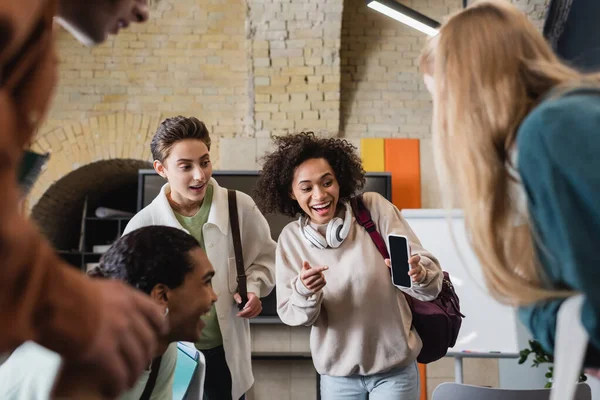 Mujer afroamericana emocionada apuntando a un teléfono inteligente cerca de amigos interracial en el aula - foto de stock