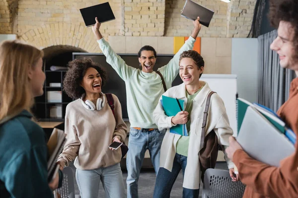 Screaming african american man holding copybooks in raised hands near interracial classmates laughing with closed eyes — Stock Photo