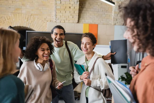 Excités camarades de classe multiethniques riant près des amis flous dans la salle de classe — Photo de stock