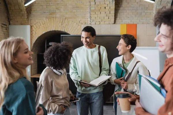 Estudiante afroamericano sonriente sosteniendo cuaderno vacío cerca de amigos multiétnicos - foto de stock