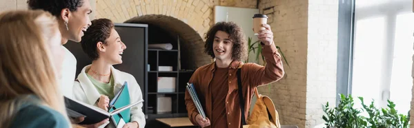 Lockiger Mann, der mit Pappbecher neben fröhlichen interrassischen Studenten anstößt, Banner — Stockfoto