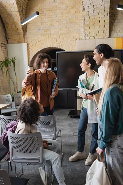 Curly man toasting with coffee to go near interracial students in auditorium — Stock Photo