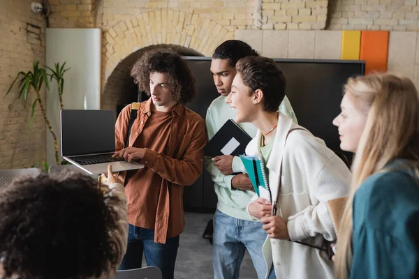 Curly man holding laptop with blank screen near friends with notebooks in university — Stock Photo