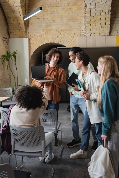 Lockiger Mann mit Laptop und leerem Bildschirm neben fröhlichen multiethnischen Klassenkameraden — Stockfoto