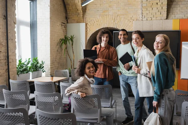 Amis multiethniques joyeux avec des cahiers et ordinateur portable souriant à la caméra dans l'auditorium — Photo de stock