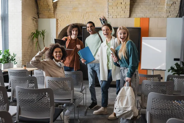 Excited multiethnic classmates shouting and showing success gesture in auditorium — Stock Photo