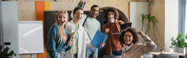 Étudiants interraciaux excités avec des copybooks montrant geste de victoire en classe, bannière — Photo de stock