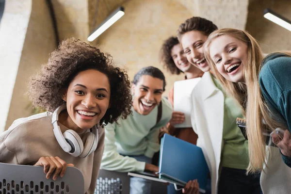 Étudiant afro-américain joyeux avec écouteurs regardant la caméra avec des amis multiculturels — Photo de stock