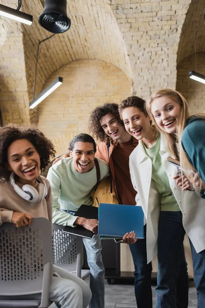 Joyful multicultural students with notebooks looking at camera in classroom — Stock Photo