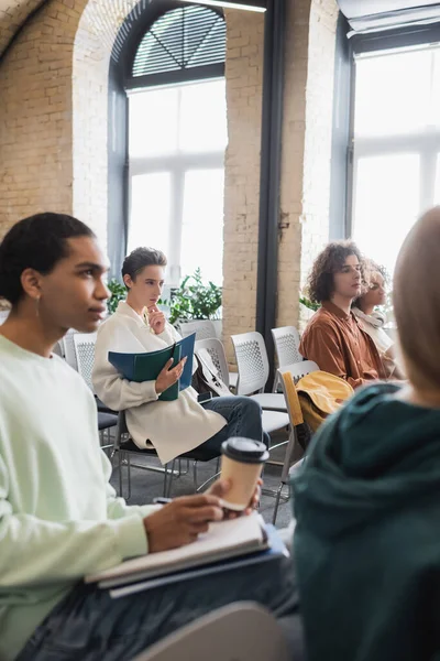 Homme afro-américain flou tenant du café pour aller tout en étant assis dans la salle de classe avec des étudiants interracial — Photo de stock