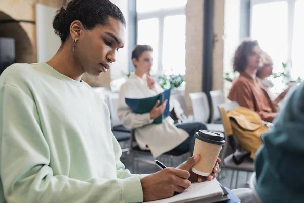 Estudante afro-americano com bebida takeaway escrevendo em notebook perto de colegas turvos — Fotografia de Stock