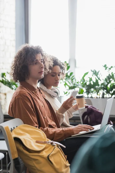 Uomo riccio utilizzando computer portatile vicino donna afro-americana con caffè per andare e telefono cellulare — Foto stock