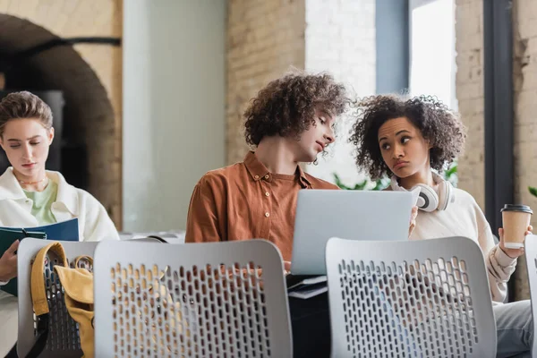 Femme afro-américaine réfléchie avec du café pour aller regarder frisé ami avec ordinateur portable dans l'auditorium — Photo de stock