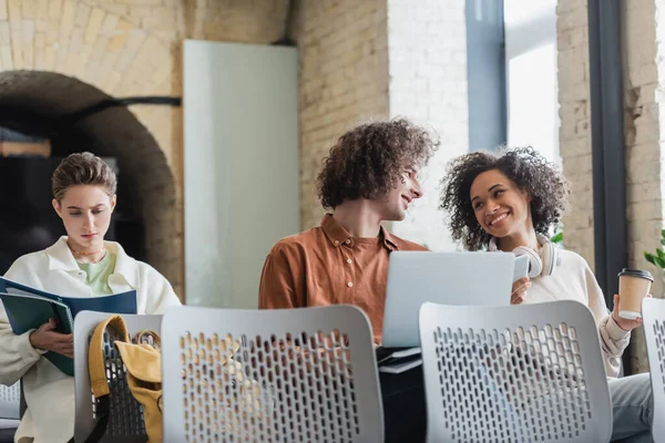 Mulher americana africana feliz com copo de papel falando com colega encaracolado com laptop no auditório — Fotografia de Stock