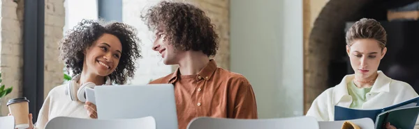 Happy african american woman with paper cup and curly student with laptop looking at each other in classroom, banner — Stock Photo