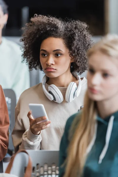 Молода афроамериканка з навушниками тримає смартфон під час лекції поблизу розмитих студентів — стокове фото