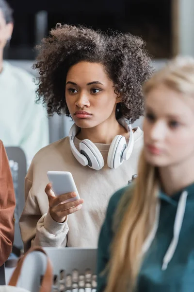 Jeune femme afro-américaine avec casque tenant smartphone pendant la conférence près des étudiants flous — Photo de stock