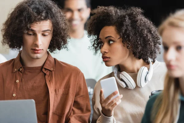 Mujer afroamericana con teléfono móvil y auriculares hablando con un amigo rizado en el aula - foto de stock