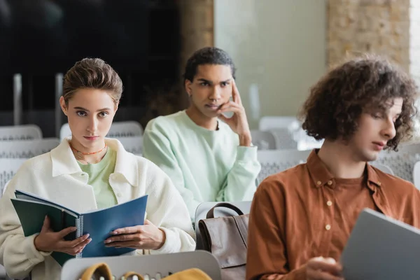 Young woman sitting with copybooks near thoughtful african american student in classroom — Stock Photo