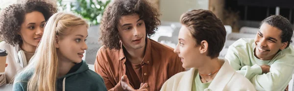 Lockiger Mann, der mit der Hand zeigt, während er mit lächelnden multiethnischen Klassenkameraden spricht, Banner — Stockfoto