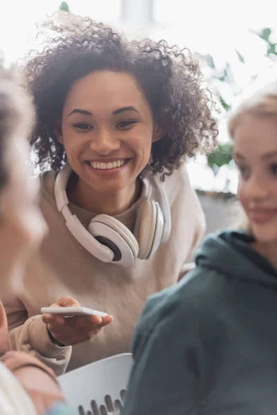 Joven afroamericana mujer con auriculares y teléfono inteligente sonriendo cerca borrosa compañeros de clase - foto de stock