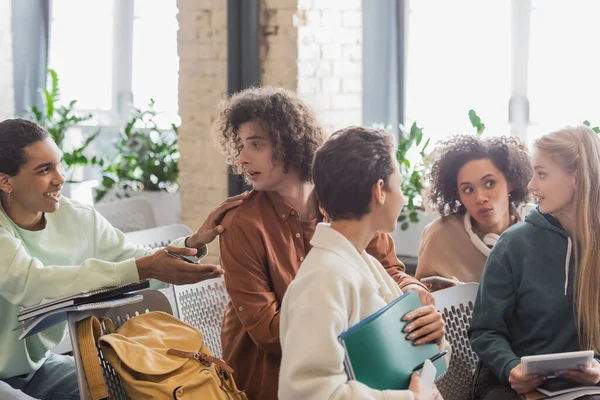 Lächelnder afrikanisch-amerikanischer Mann, der die Schulter eines Freundes berührt, während er mit ihm in der Nähe interrassischer Klassenkameraden spricht — Stockfoto