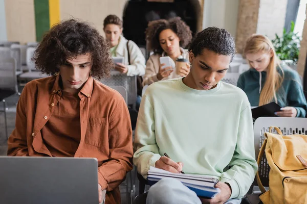 Studente africano americano che scrive nel taccuino vicino al compagno di classe riccio con il computer portatile — Foto stock