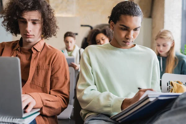 Afrikanisch-amerikanischer Student schreibt in Notizbuch, während Freund während des Unterrichts auf Laptop tippt — Stockfoto