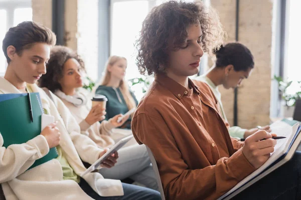 Homme bouclé écrit dans un cahier pendant la conférence près des étudiants multiethniques — Photo de stock