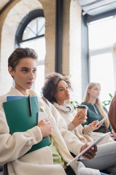 Young woman with copybooks and gadgets listening lecture near interracial classmates — Stock Photo