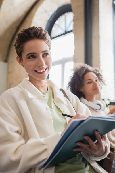 Donna sorridente che scrive nel taccuino vicino al compagno di classe afro-americano su sfondo sfocato — Foto stock