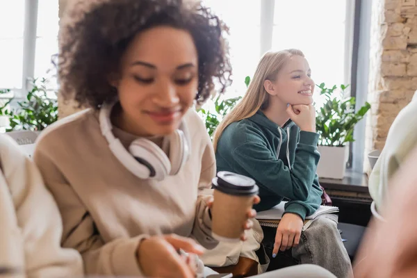 Estudiante sonriente escuchando conferencia cerca borrosa compañero de clase afroamericano con auriculares y café para llevar - foto de stock