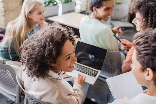 Cheerful african american student with laptop looking to blurred friend in classroom — Stock Photo