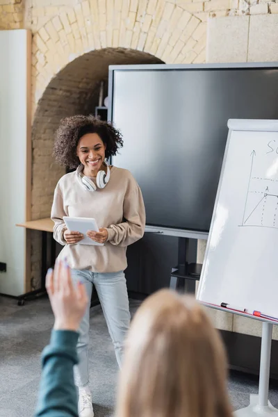 Cheerful african american student with digital tablet near whiteboard with task and blurred classmate with raised hand — Stock Photo